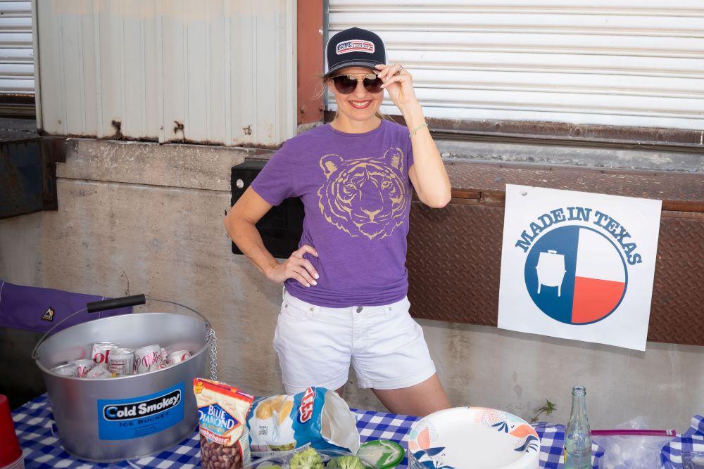 Person wearing the American Flag Old Smokey logo hat with a purple shirt, white shorts, and sunglasses, standing next to a table with snacks and a 'Made in Texas' sign.