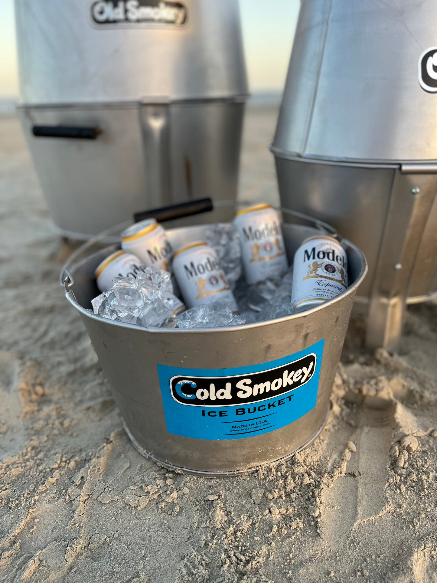 Metal ice bucket with Cold Smokey branding on a sandy surface with beer cans and ice.